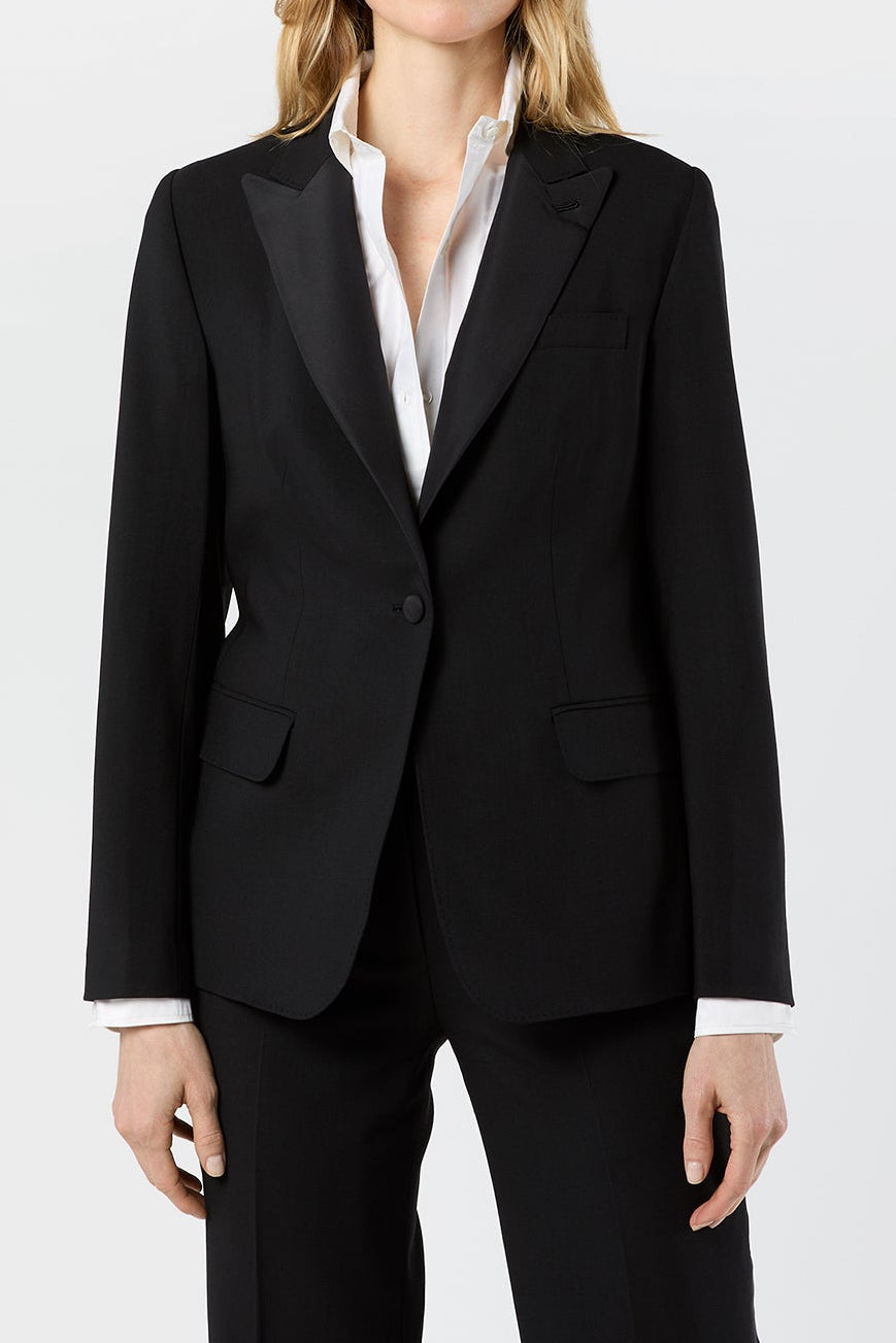 A woman modeling a black tailored blazer with black satin lapels, paired with black trousers and a white collared shirt, against a plain light gray studio background.
