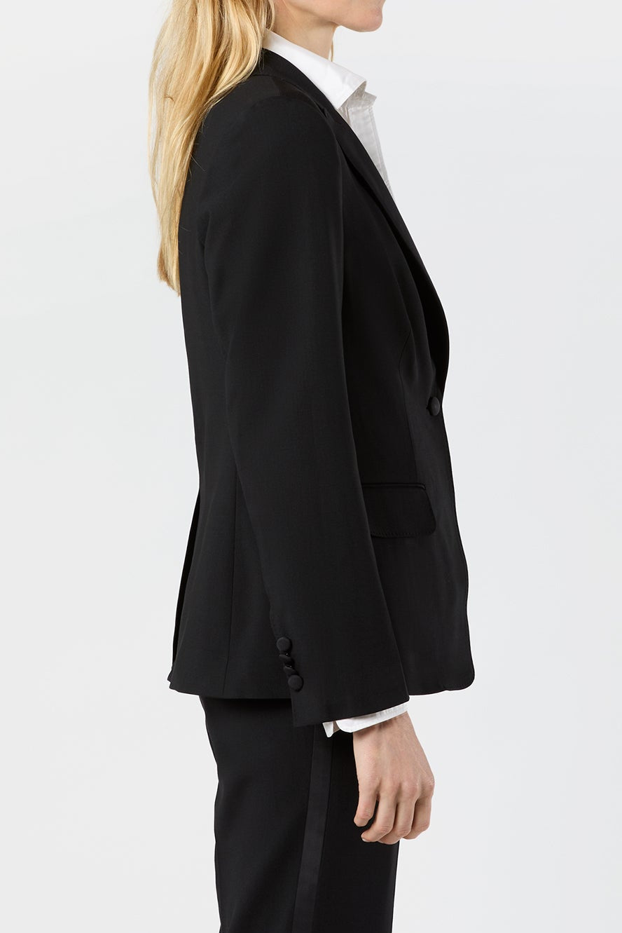 A woman in a black tailored blazer and matching trousers, styled with a white collared shirt, shown in profile against a plain white studio background.