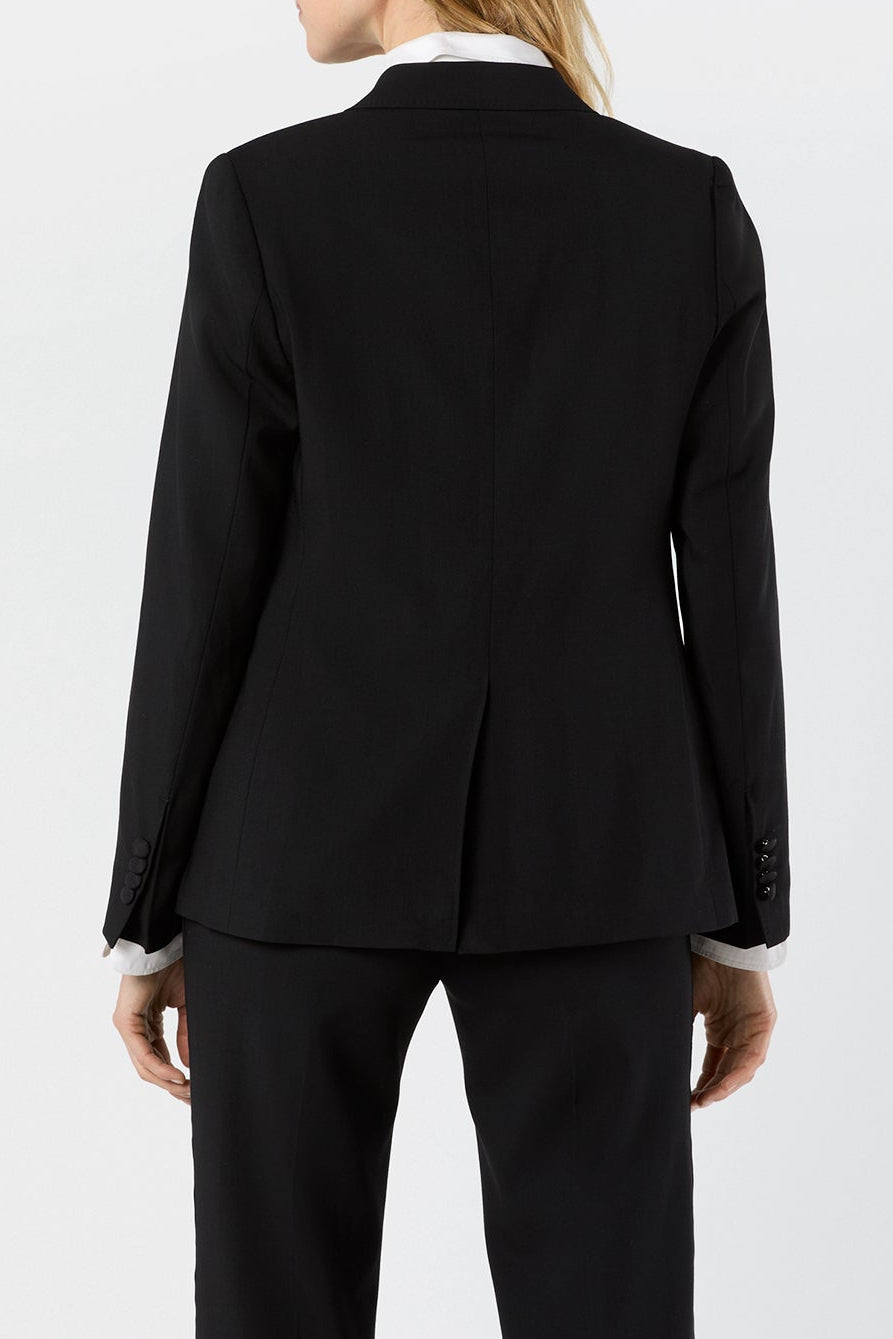 A woman shown from the back wearing a black tailored blazer and matching trousers over a white collared shirt, photographed against a clean light gray studio background.