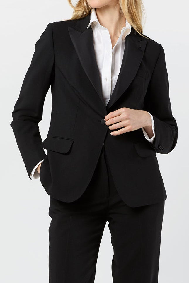 A woman modeling a black tailored pantsuit with a blazer and matching trousers, worn over a white collared shirt and black heels, against a plain light gray studio background.