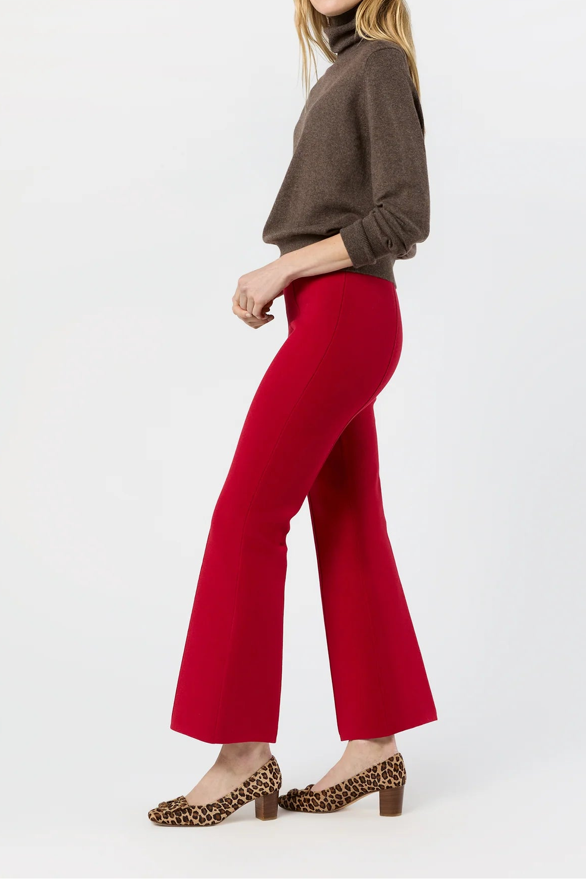 A woman modeling bright red wide-leg cropped pants with a brown turtleneck sweater and leopard-print heels, shown in profile against a plain white studio background.