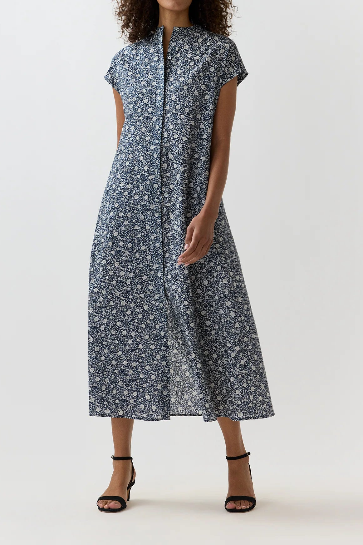A woman modeling a short-sleeve midi dress with a small white floral print on a slate blue-gray background, paired with black strappy heels, against a light gray studio backdrop.