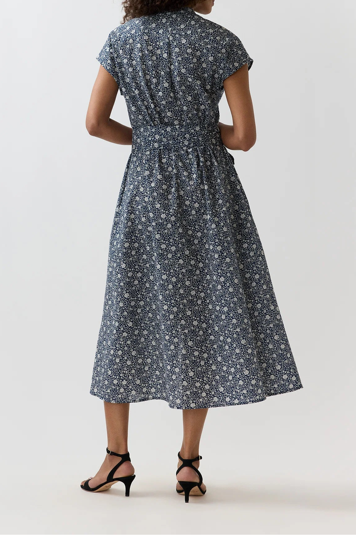 A woman viewed from the back wearing a short-sleeve midi dress with a small white floral print on a slate blue-gray background, paired with black strappy heels, against a plain light gray studio backdrop.
