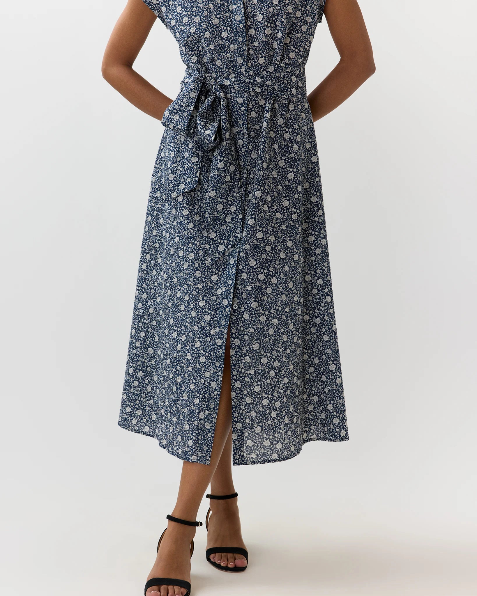 A woman wearing a sleeveless midi dress with a small white floral print on a navy blue background, featuring a tie waist and a front slit, paired with black strappy high-heeled sandals.
