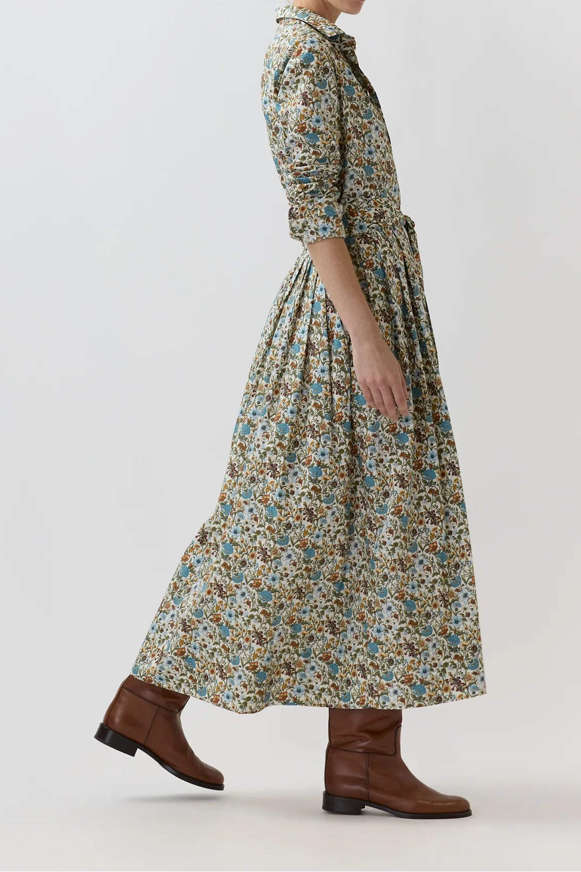 A woman modeling a long-sleeved, knee-length floral dress with a tie-neck and waist, paired with brown leather ankle boots, set against a plain light gray studio background.