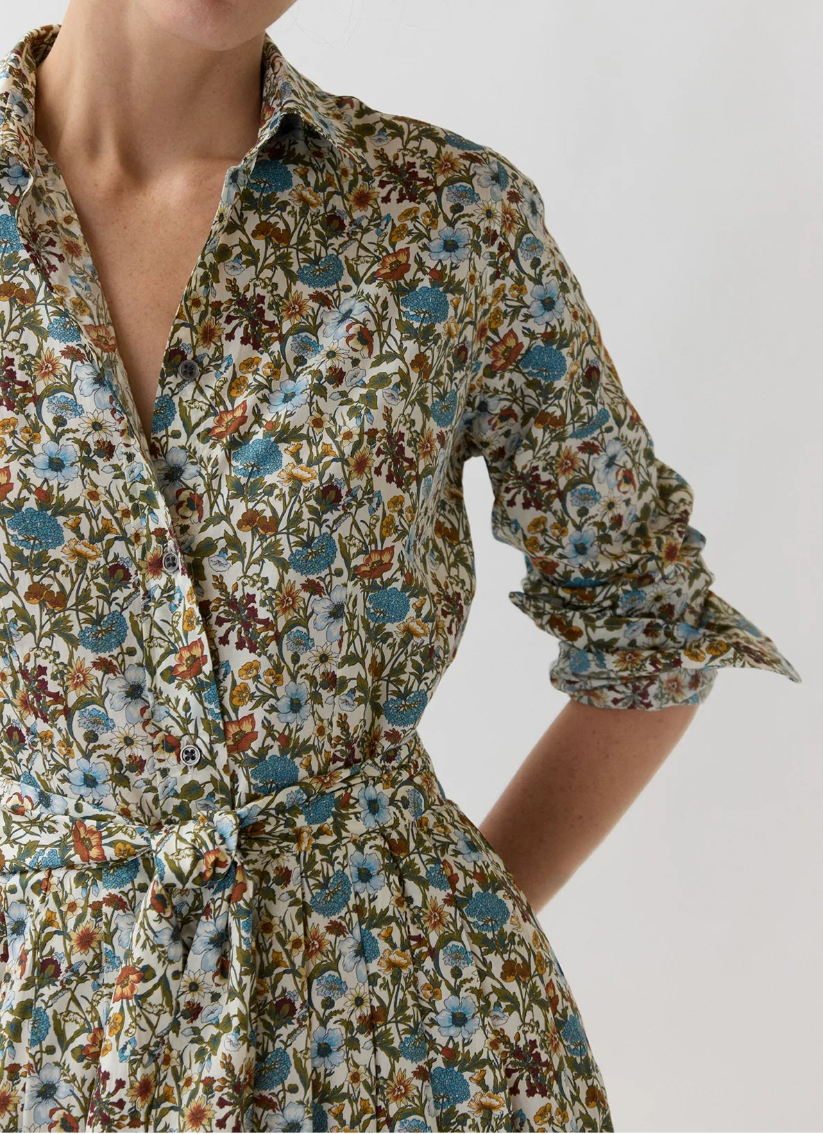 A woman wearing a floral print shirt dress with a tie waist and rolled sleeves, photographed against a plain light gray studio background.