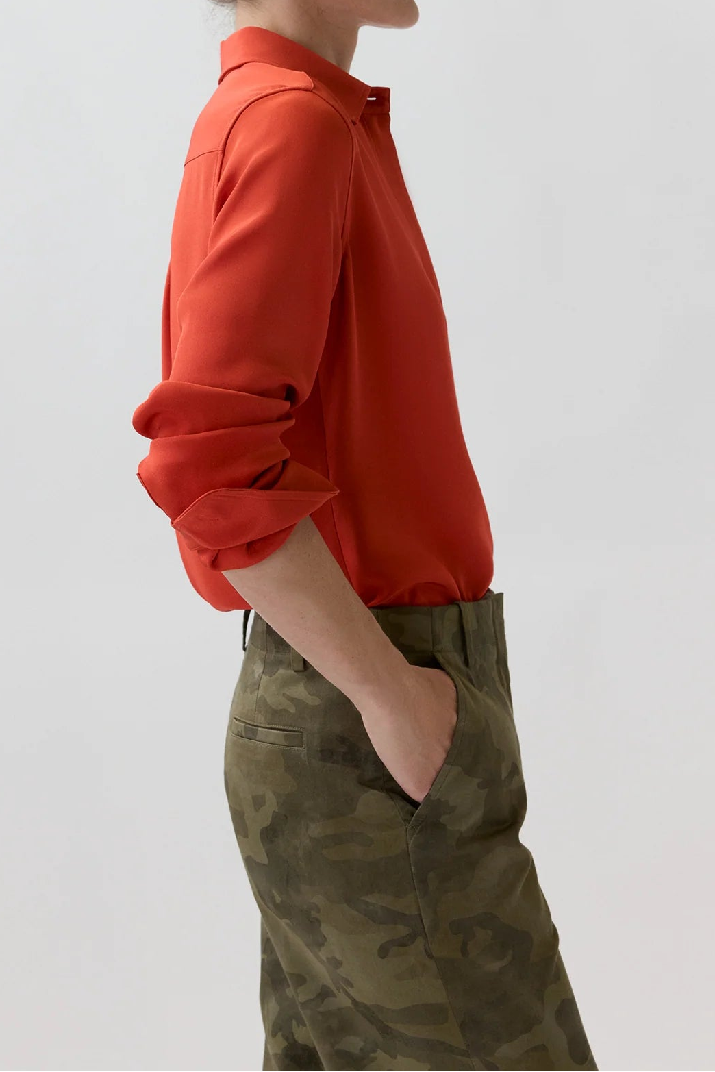 A woman in profile wearing a bright orange-red long-sleeve button-up shirt and olive green camouflage trousers, standing against a plain light gray studio background.