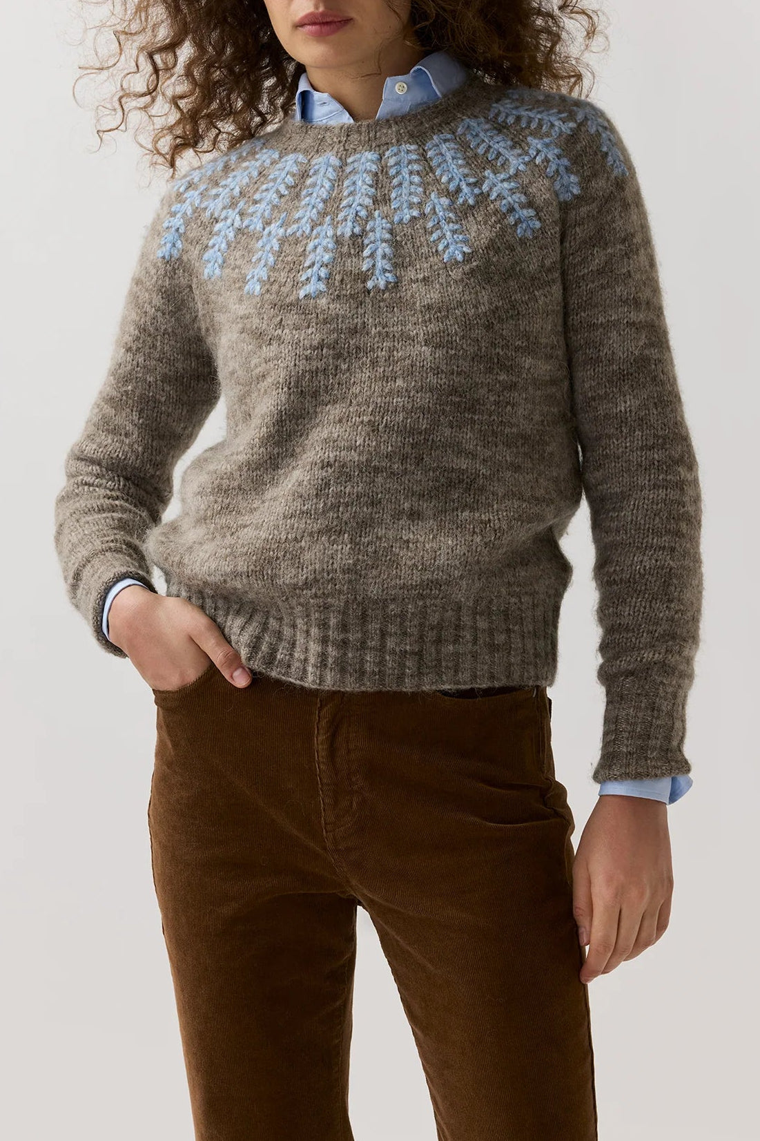 A woman modeling a brown knit sweater with a light blue and white Fair Isle yoke pattern, worn over a light blue collared shirt and brown corduroy pants, against a plain light gray background.