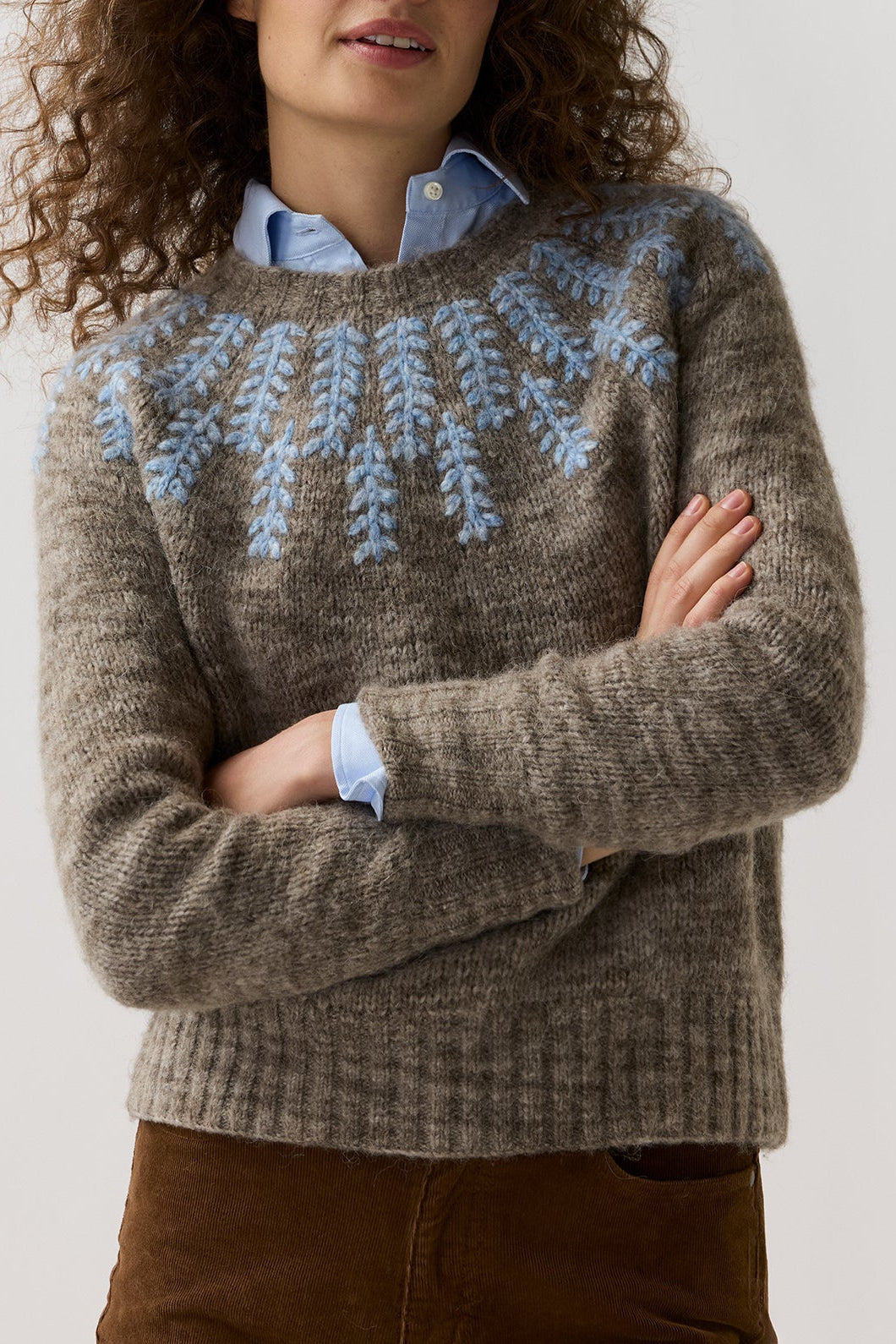 A woman with curly brown hair wearing a brown knit sweater with a light blue Nordic-style pattern across the chest, layered over a light blue collared shirt and brown trousers, against a plain light gray background.