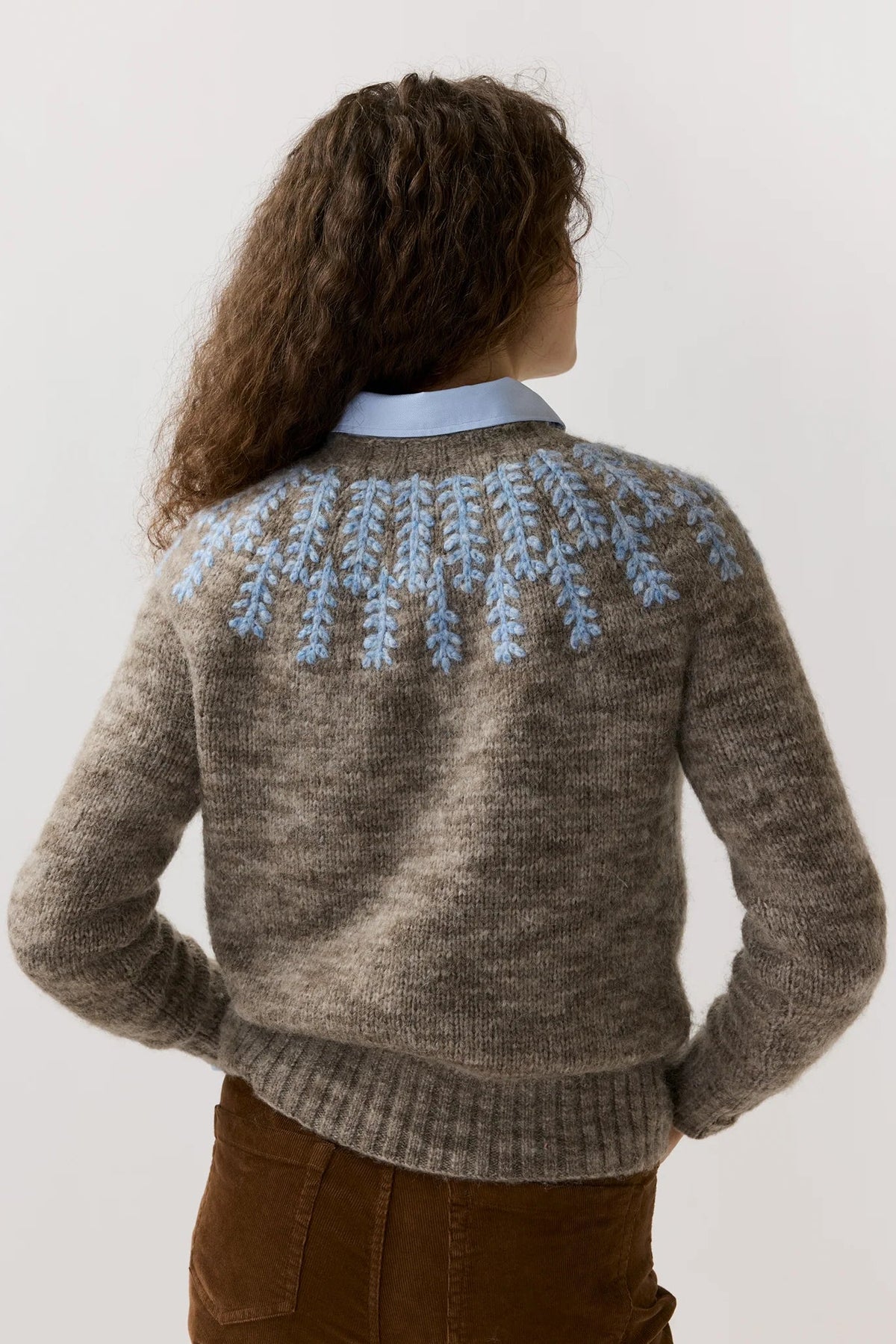 A woman viewed from behind wearing a brown knit sweater with a light blue embroidered leaf pattern across the shoulders, layered over a light blue collared shirt and brown pants, against a plain light gray background.