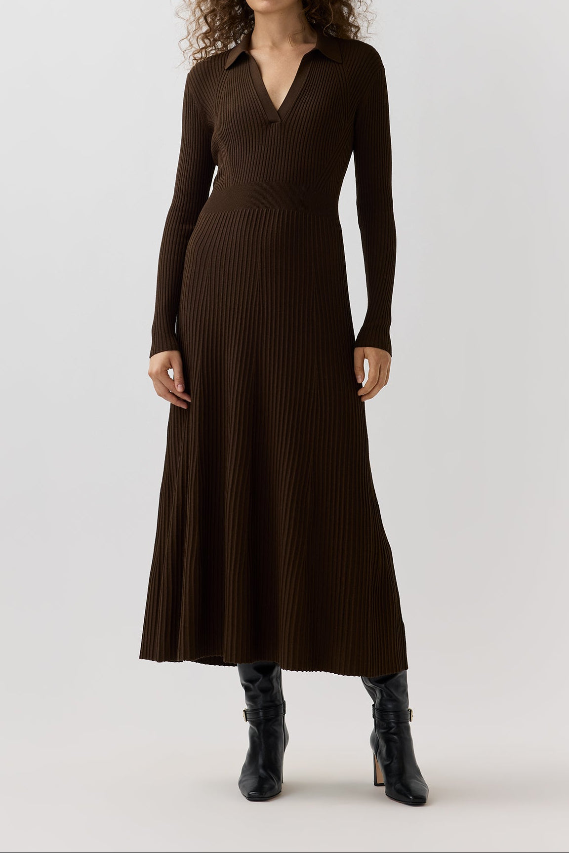 A woman modeling a dark brown ribbed knit midi dress with long sleeves and a V-neckline, paired with black ankle boots, against a plain light gray studio background.