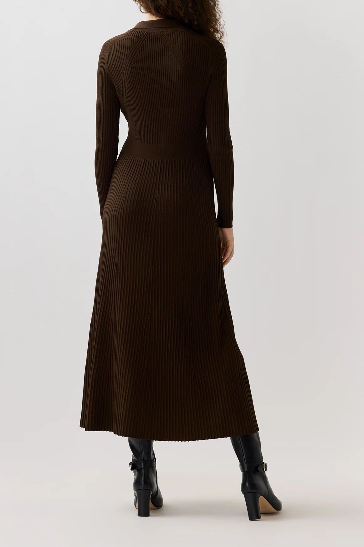 A woman viewed from behind wearing a dark brown ribbed long-sleeve midi dress and black ankle boots against a light gray studio background.