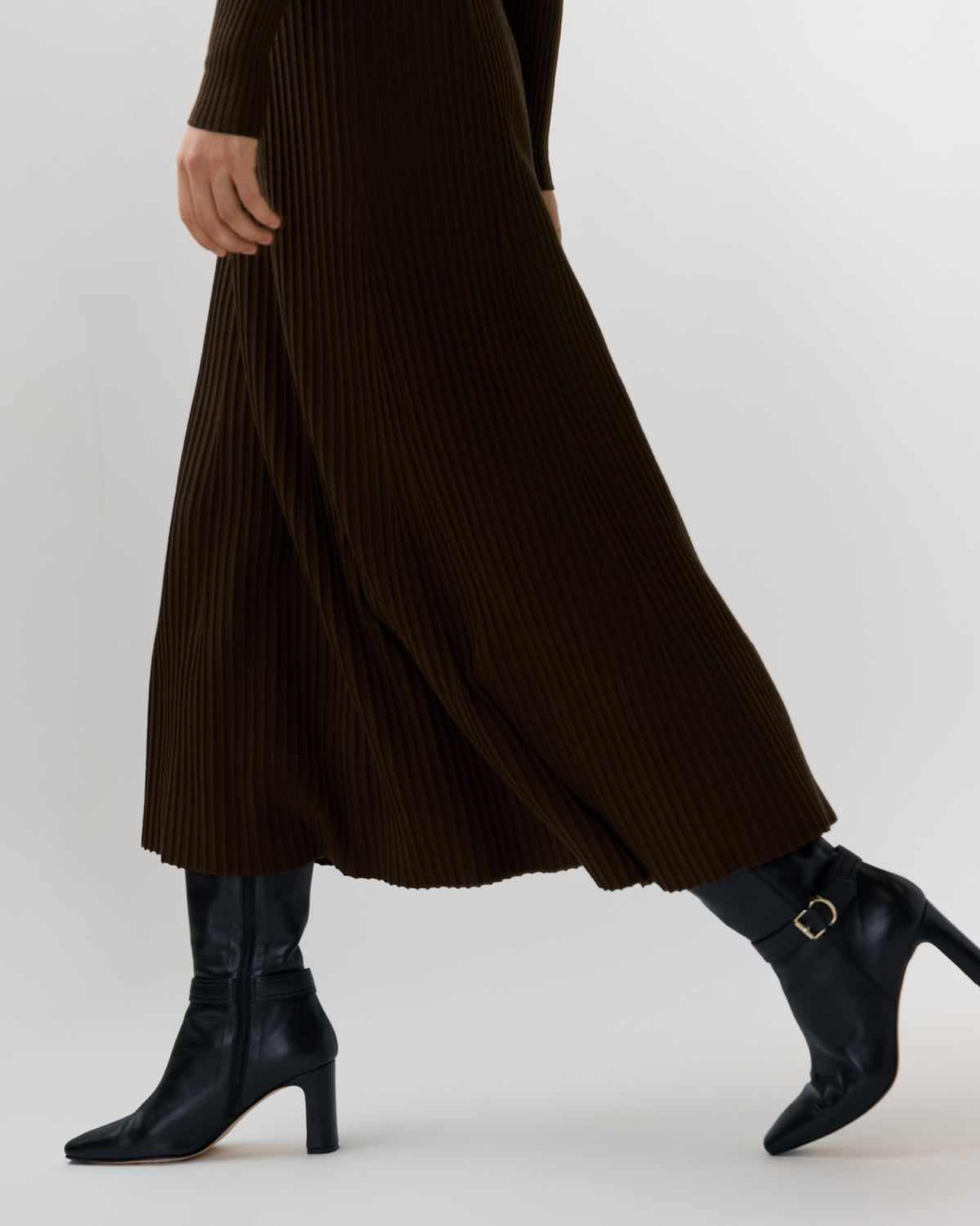 A person wearing a dark brown pleated maxi skirt and black ankle boots with gold buckle details, shown from the waist down against a light gray studio background.