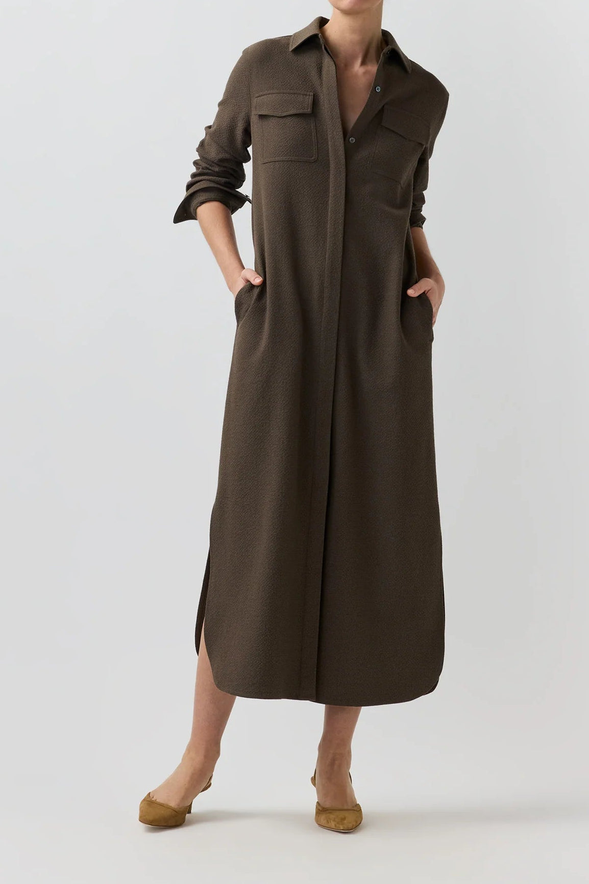 A model wearing a long, dark brown button-up shirt dress with front flap pockets and side slits, paired with tan flat shoes, against a light gray studio background.