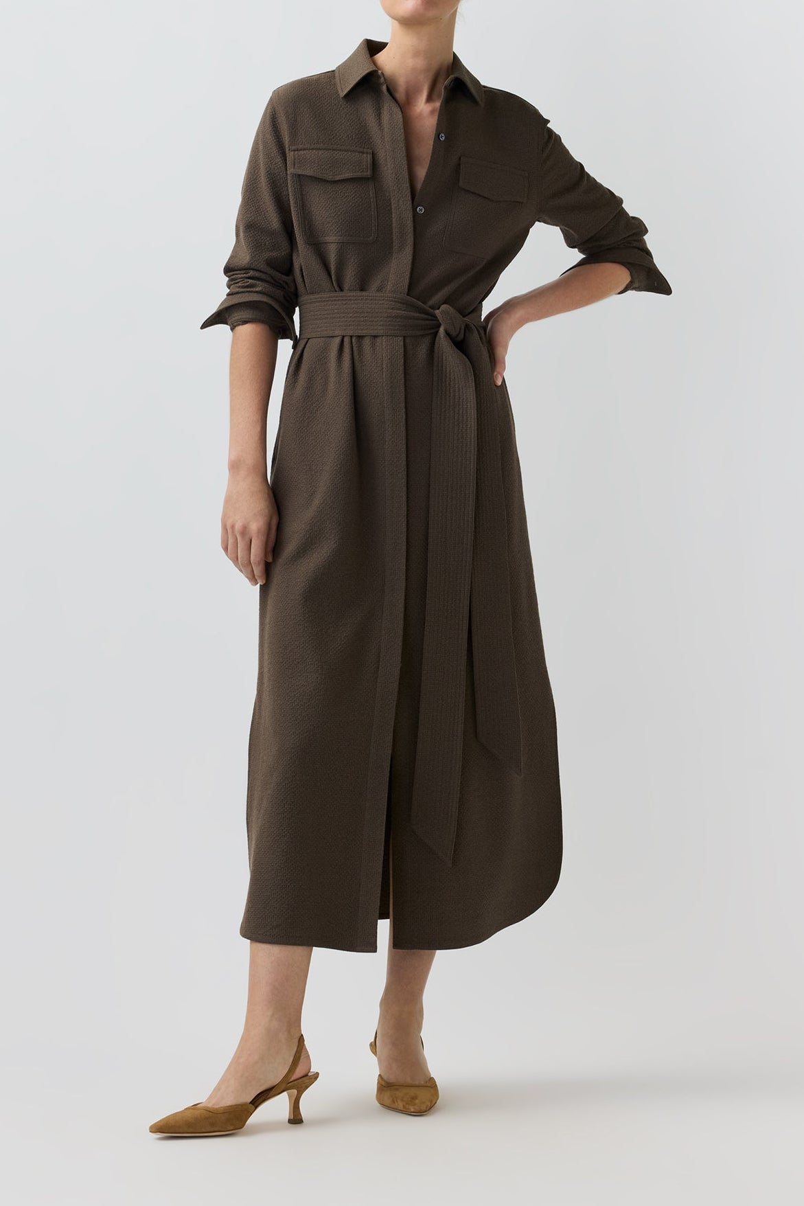 A woman modeling a dark brown midi shirt dress with a tie waist belt, three-quarter sleeves, and chest pockets, paired with tan heeled sandals against a light gray studio background.
