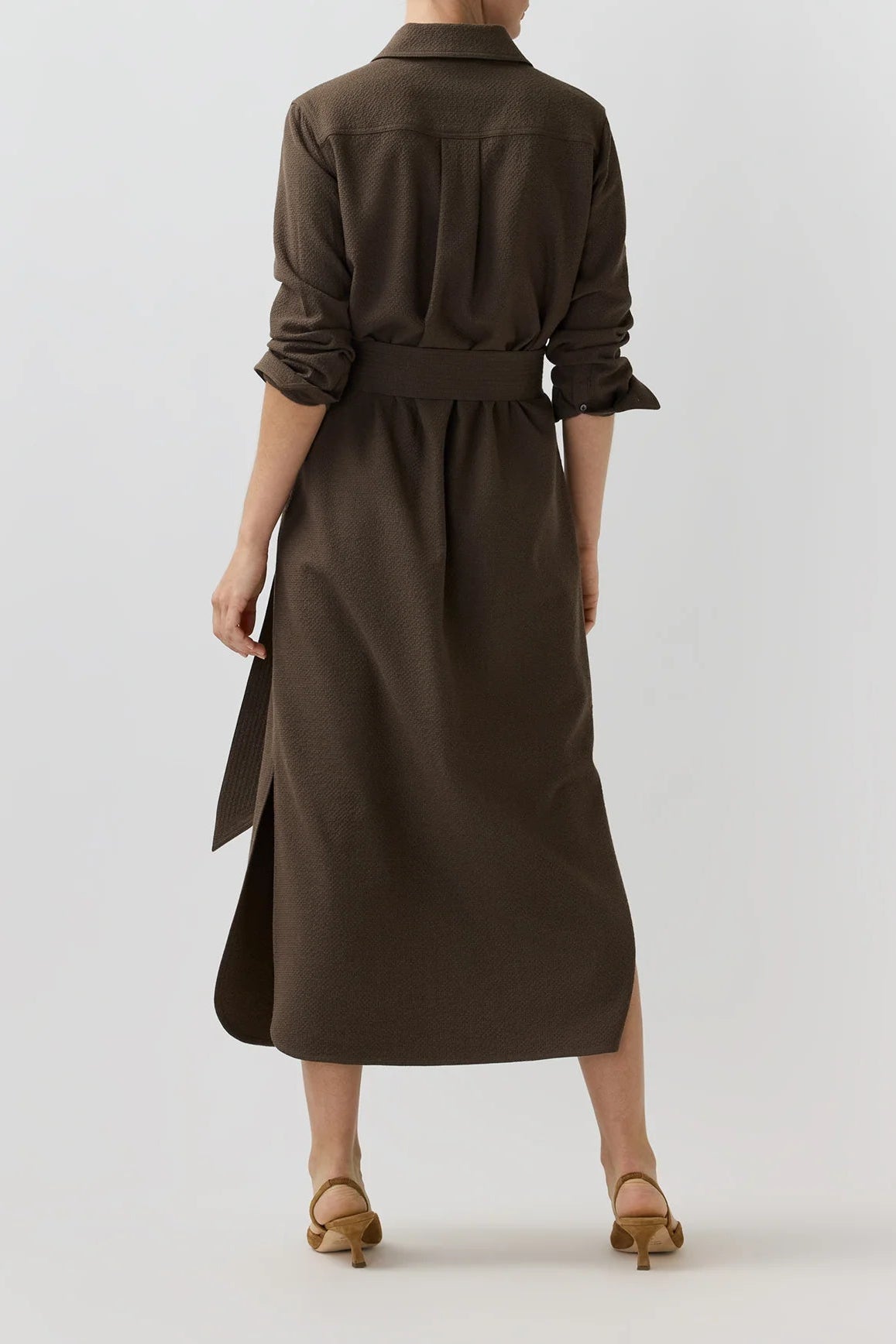 A woman viewed from the back wearing a dark brown midi shirt dress with a waist tie and rolled sleeves, paired with tan heeled sandals, against a light gray studio background.