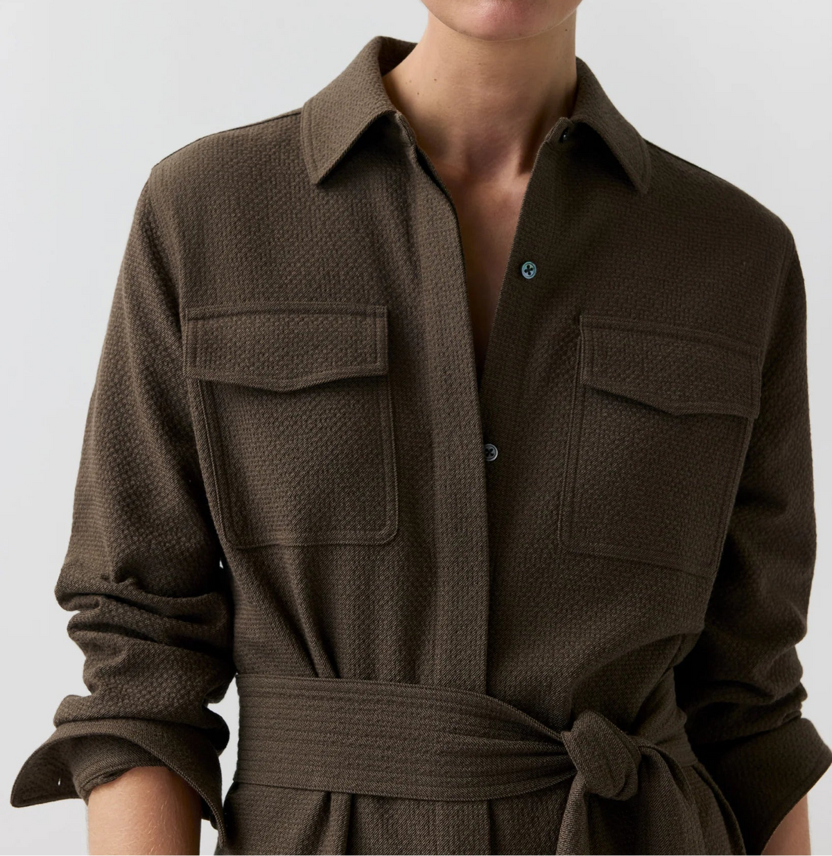 A close-up of a model wearing a dark brown textured shirt dress with a collar, button front, chest pockets, and a matching tie belt.