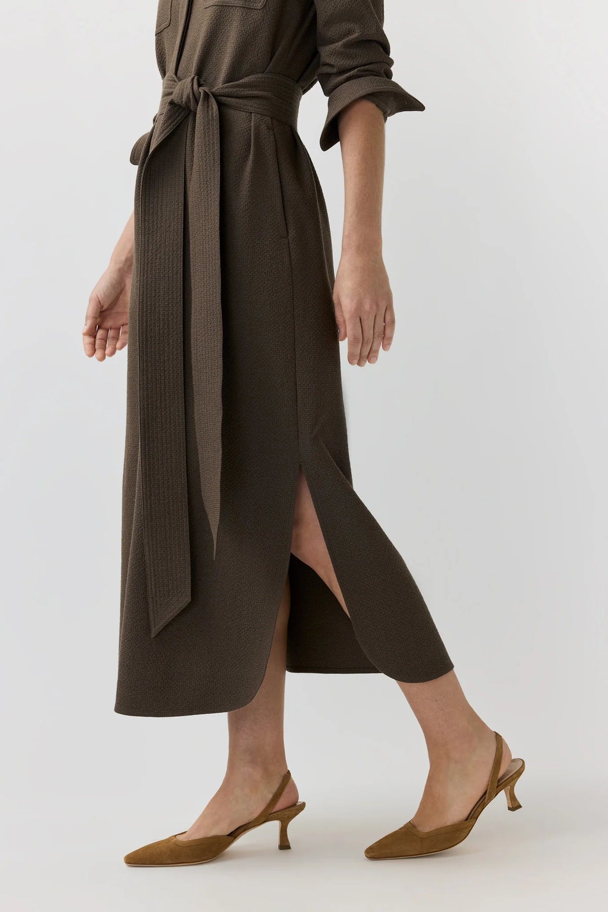A model wearing a dark brown midi wrap dress with a tie waist and side slit, paired with brown slingback kitten heels, against a light gray studio background.