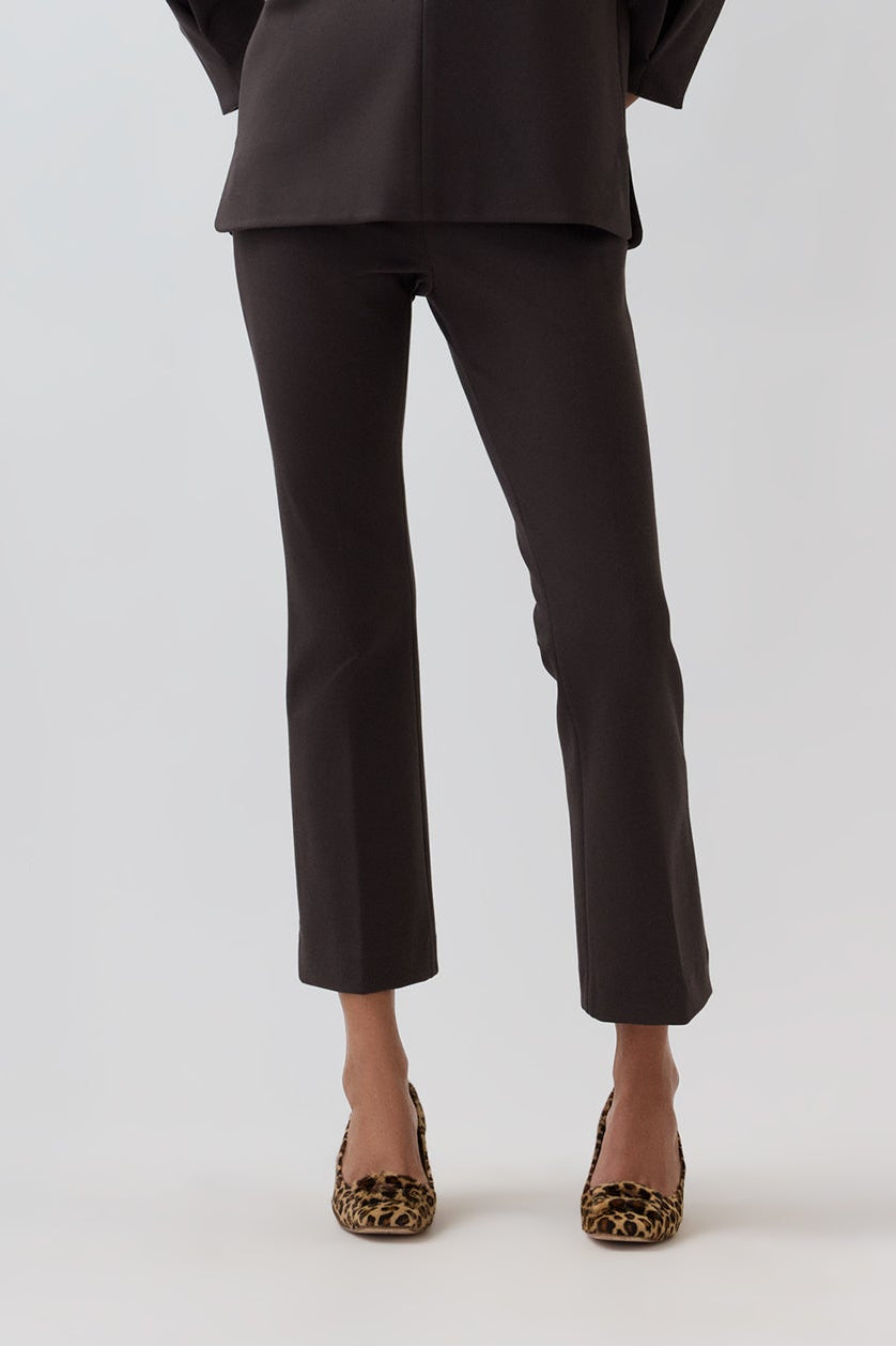 A model with curly hair stands against a light gray studio background wearing a dark brown long-sleeve collared jacket and matching cropped trousers, paired with leopard-print heels.
