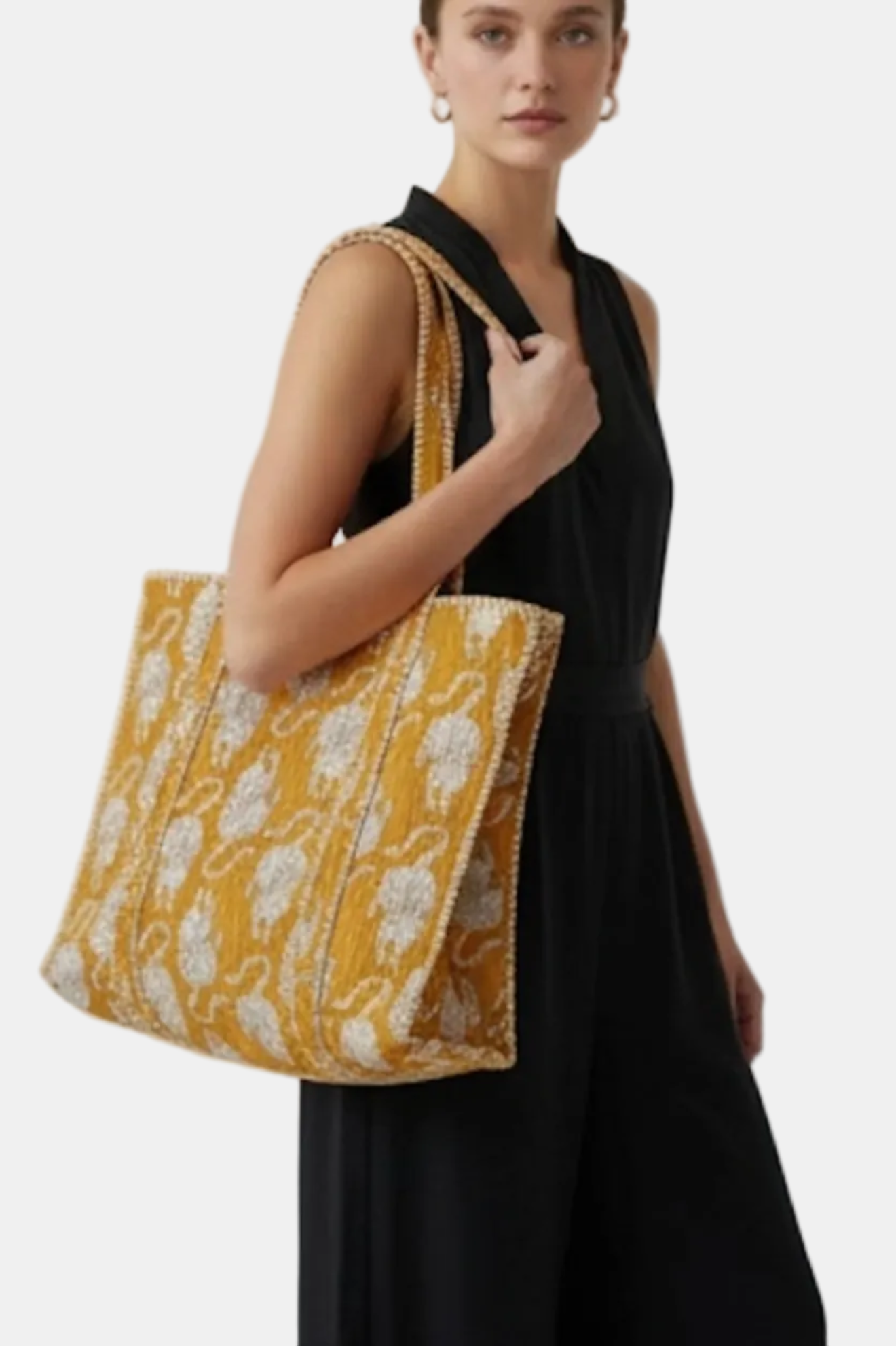 A woman wearing a black sleeveless dress carries a large mustard yellow and white patterned tote bag with gold-toned chain handles against a plain light gray background.