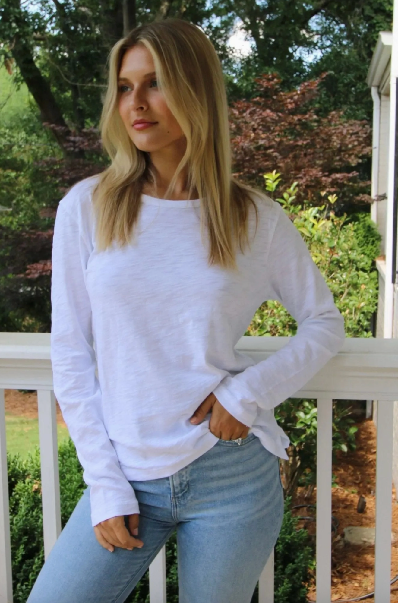 A woman wearing a white long-sleeve crewneck t-shirt and light blue jeans, standing on a white porch railing with a garden background.