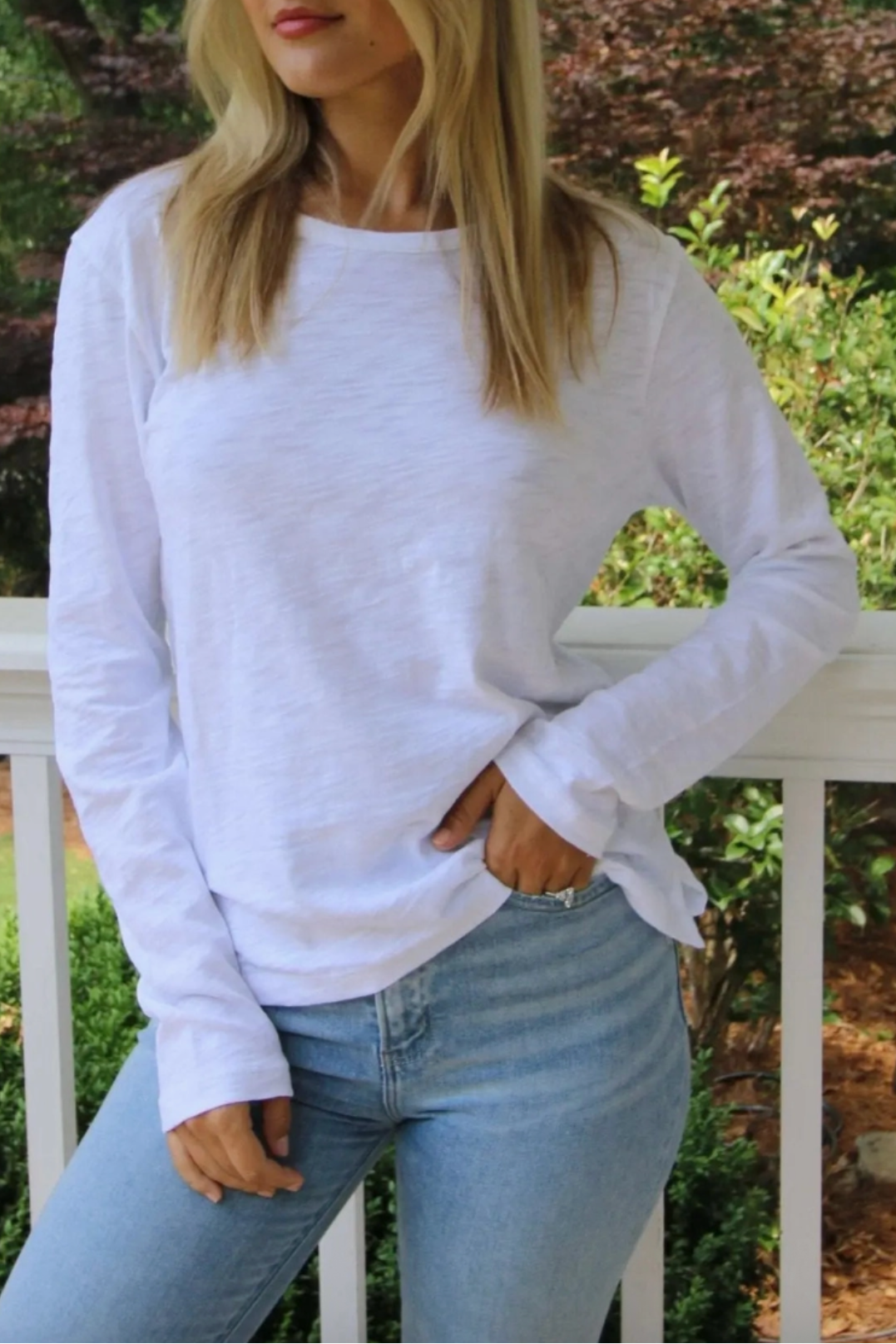 A woman wearing a white long-sleeve crewneck t-shirt and light blue jeans, standing on a white porch railing with a garden background.