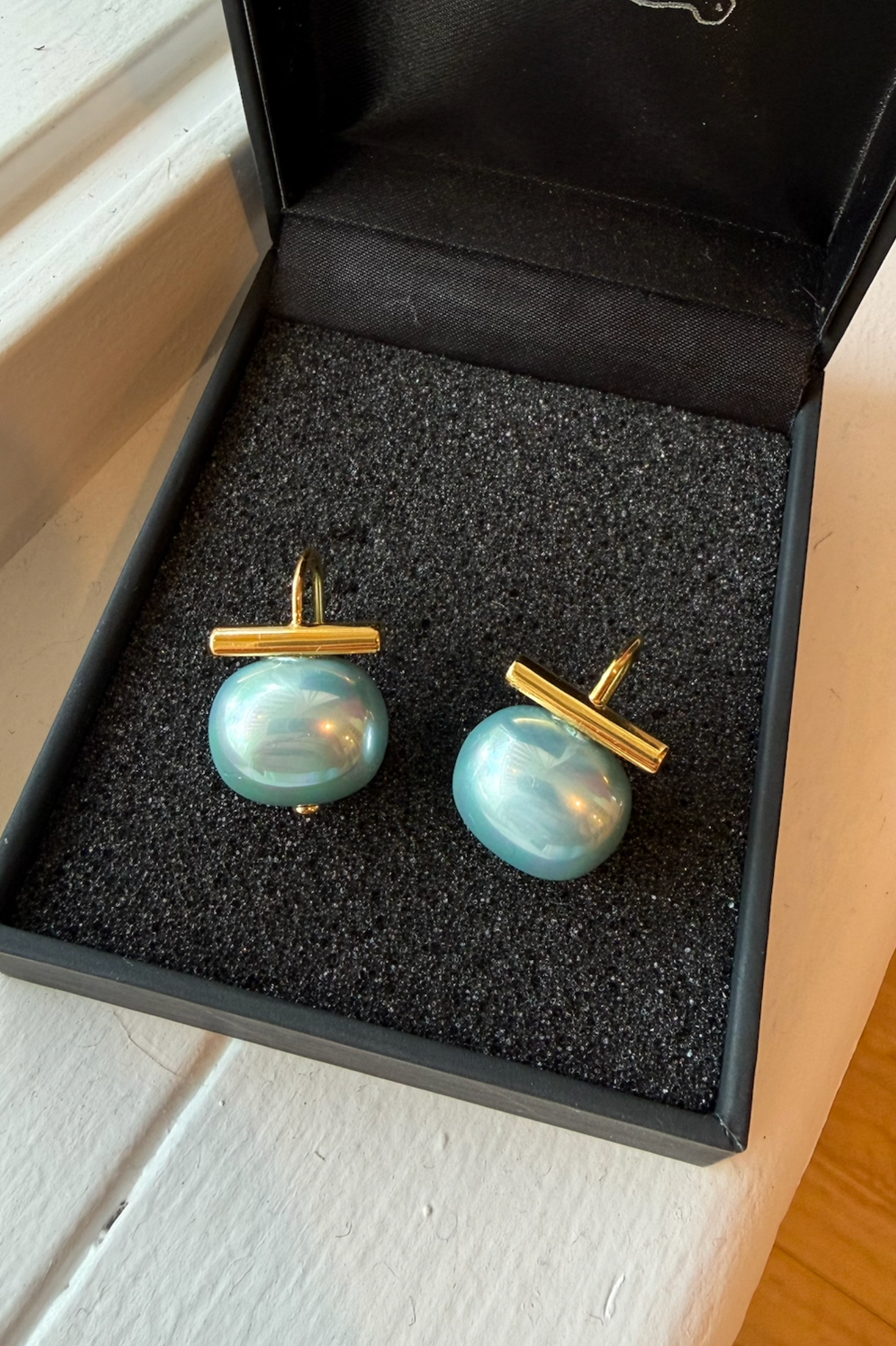 A pair of gold T-bar cufflinks with round light blue-green freshwater pearl backings, presented inside an open black jewelry box.