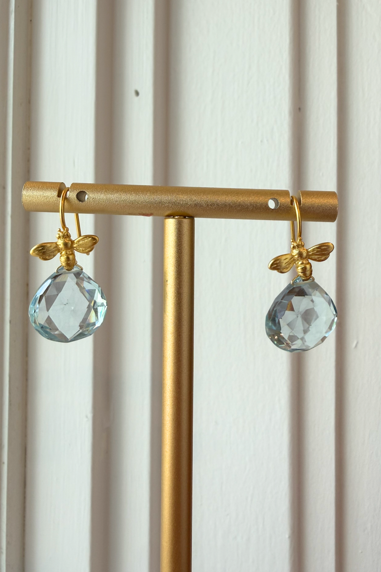 A pair of gold-tone drop earrings with faceted light blue teardrop crystal pendants, hanging from a gold display stand against a white paneled background.