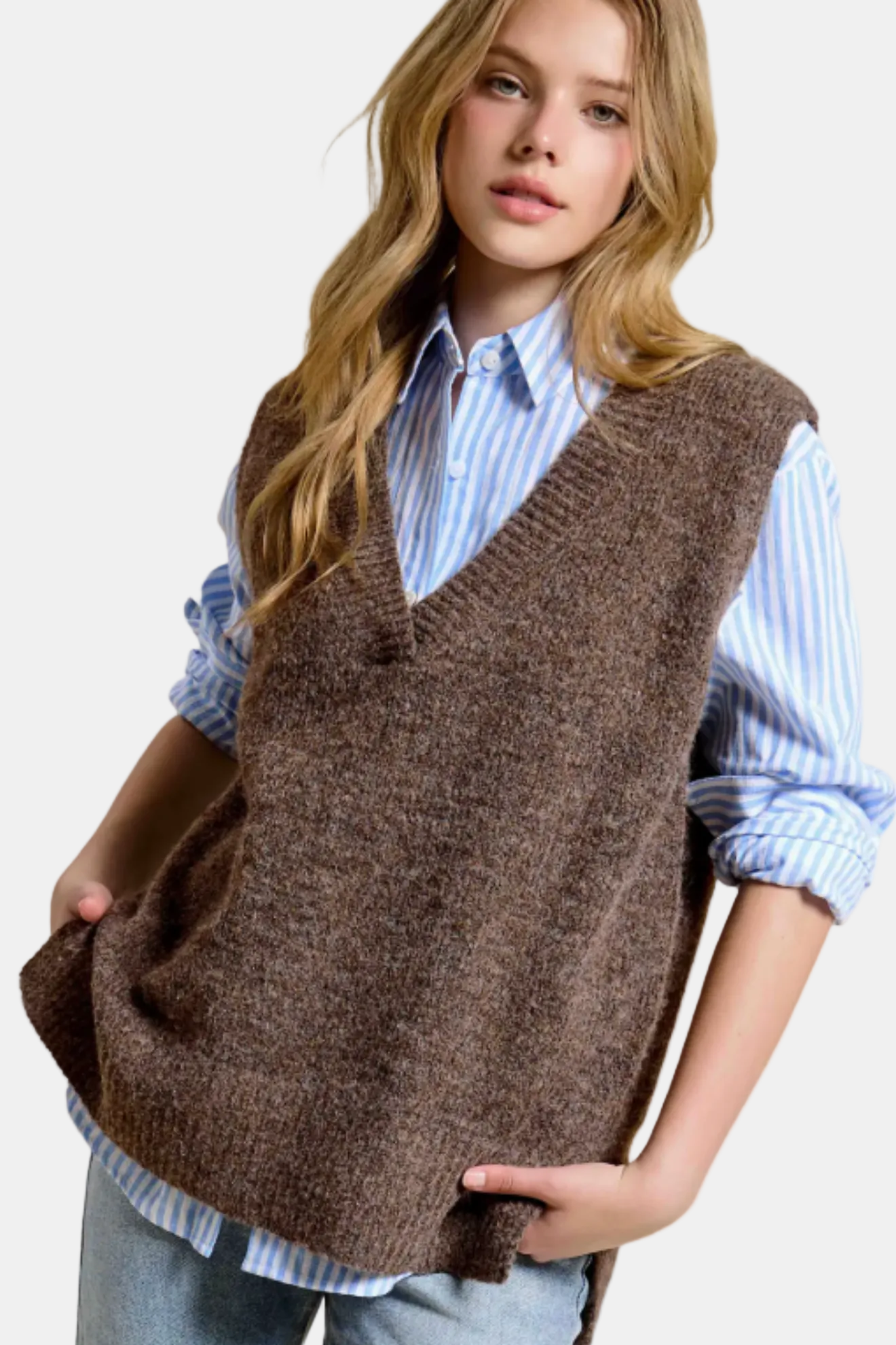 A young woman wearing a brown knit sweater vest over a light blue and white striped button-down shirt, paired with light-wash jeans, against a plain white background.