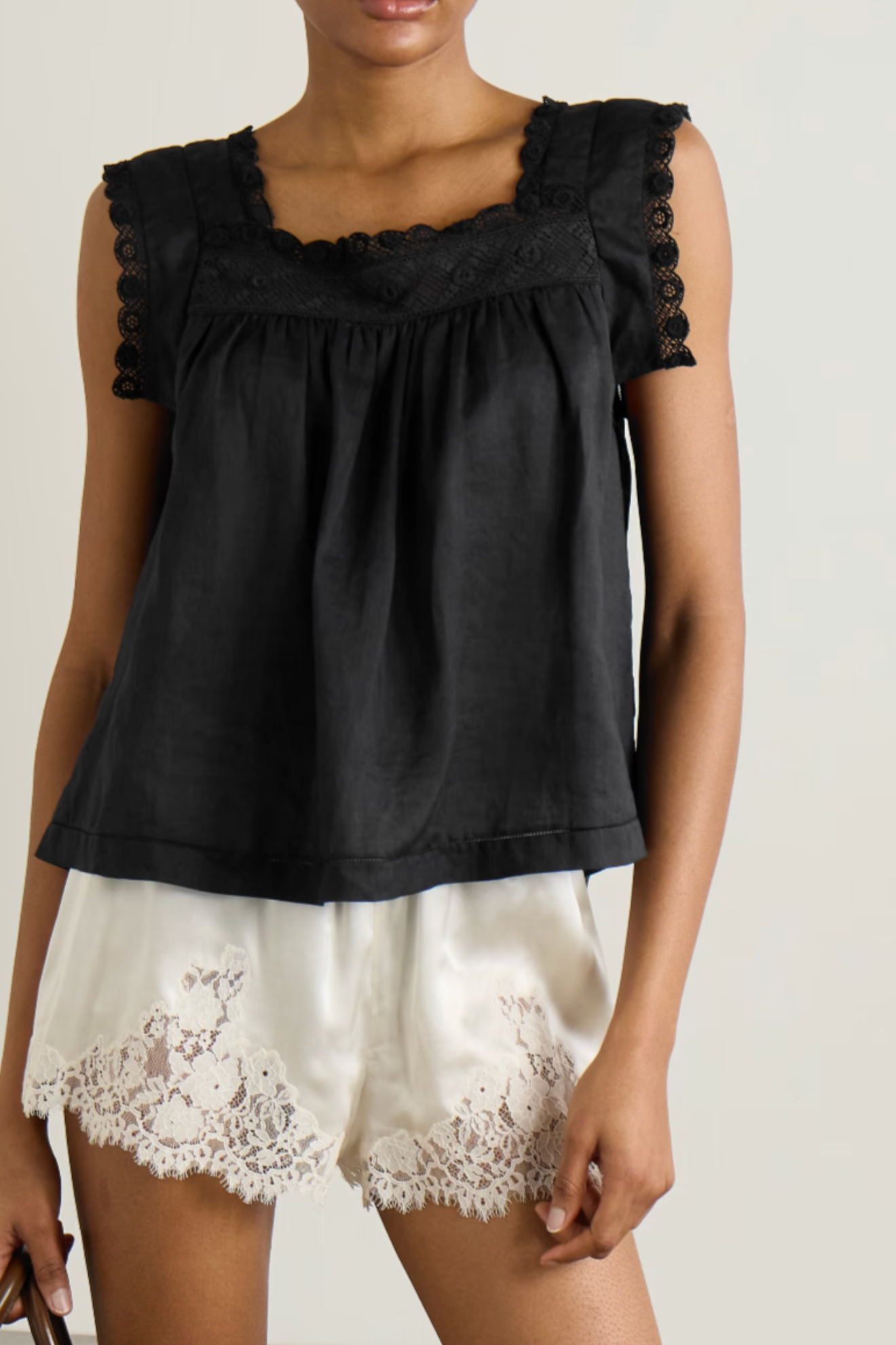 A woman wearing a black sleeveless top with a square neckline, scalloped lace trim, and gathered shoulders, paired with white lace-trimmed shorts against a plain light background.