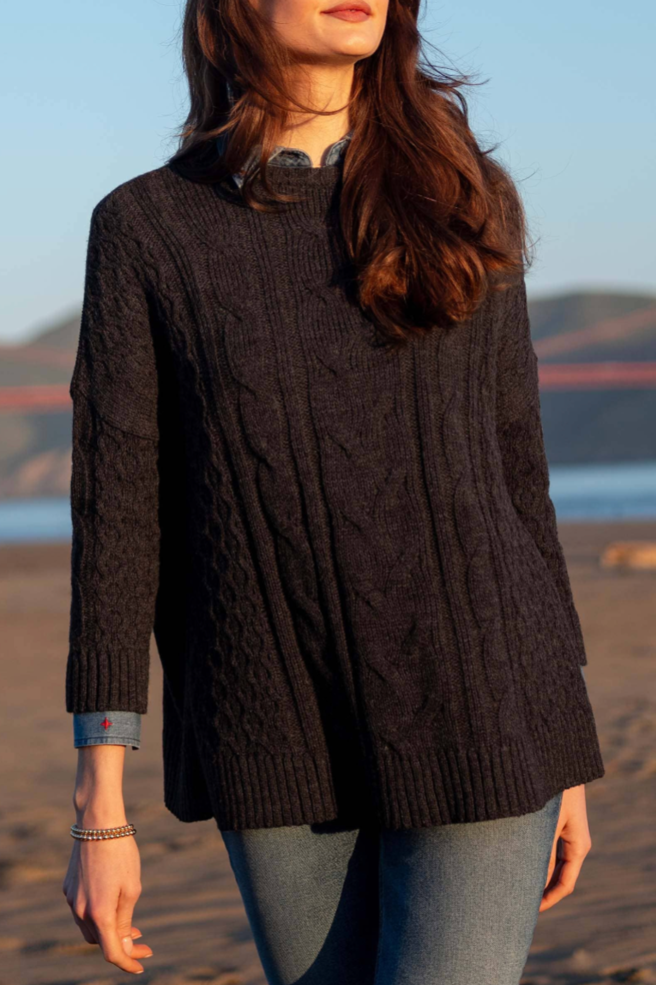 Person wearing a dark brown cable knit sweater on a beach with mountains in the background