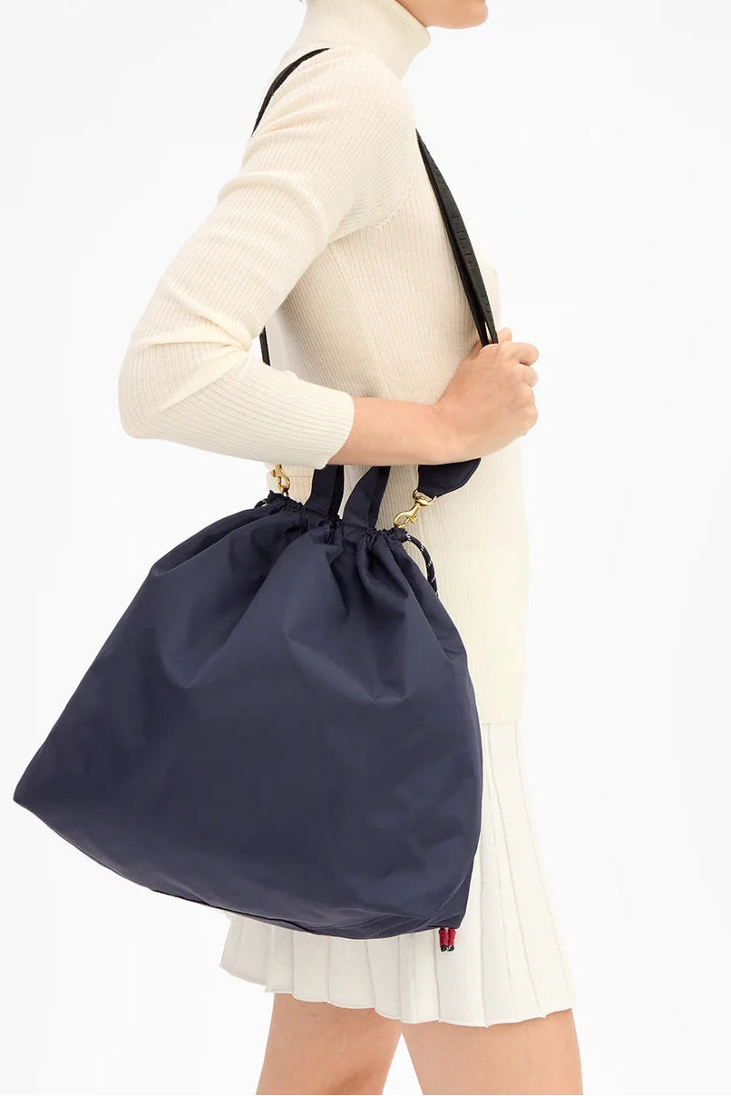 A woman in a cream ribbed turtleneck sweater and white pleated skirt shown in profile, carrying a large navy drawstring bag over her shoulder against a plain white studio background.
