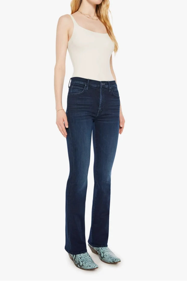 A woman modeling dark blue flare jeans with a cream ribbed tank top and patterned green shoes, standing against a plain white background.