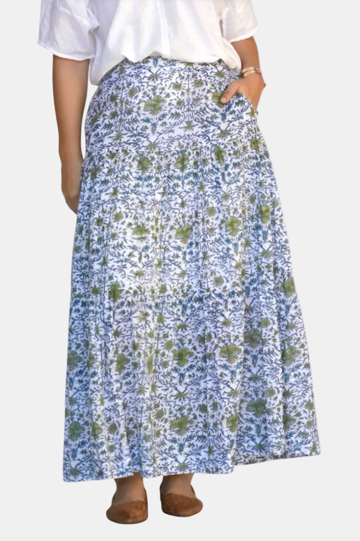 A woman wearing a long printed maxi skirt with a blue and green floral pattern, paired with a white top and brown flat shoes, shown against a plain white background.