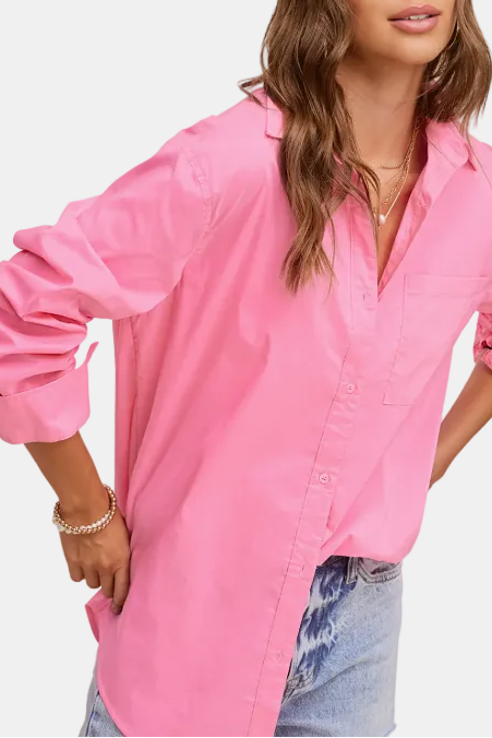 A woman wearing a bright pink button-up shirt with rolled-up sleeves and light-wash blue jeans, posed against a plain light beige background.