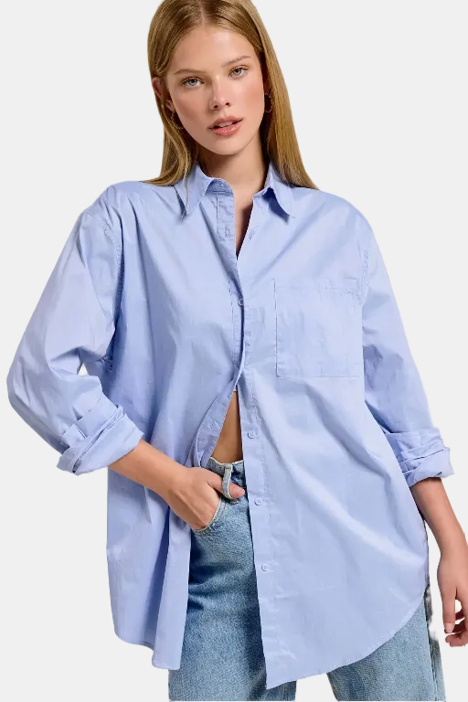 A woman wearing an oversized light blue button-up shirt with rolled sleeves and light-wash jeans, standing indoors near large windows.