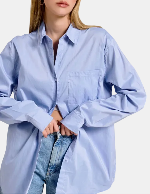 A woman wearing an oversized light blue button-up shirt with a chest pocket, paired with blue jeans, shown against a light cream background with a subtle leaf motif on the right.