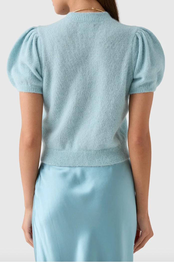A woman shown from the back wearing a light blue knit short-sleeve top with puffed sleeves and a matching light blue satin skirt, against a plain light gray studio background.
