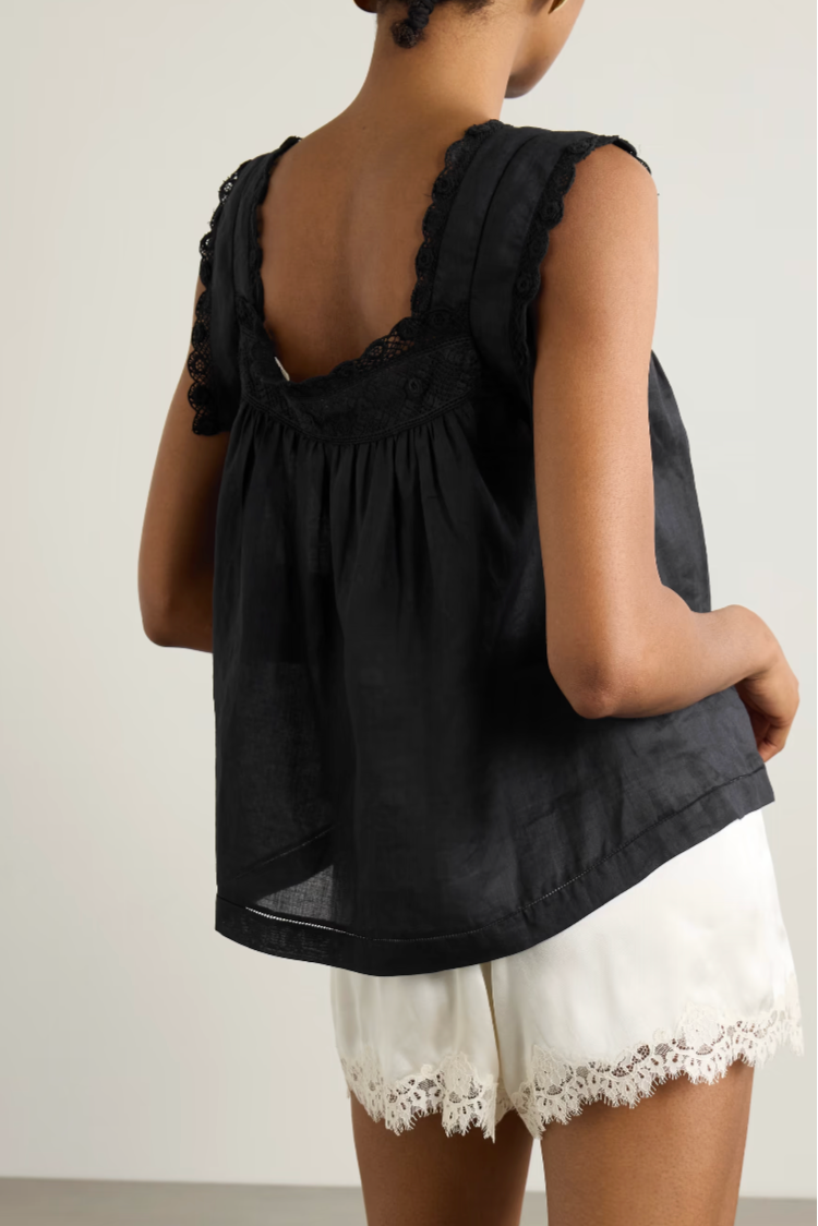 A woman shown from the back wearing a black sleeveless top with ruffled trim and a gathered back, paired with white shorts featuring lace trim, against a plain light gray studio background.
