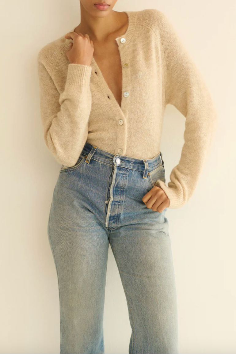 A woman modeling a light beige open-front cardigan with buttons, worn over a matching beige top and paired with light-wash blue jeans, against a plain off-white background.