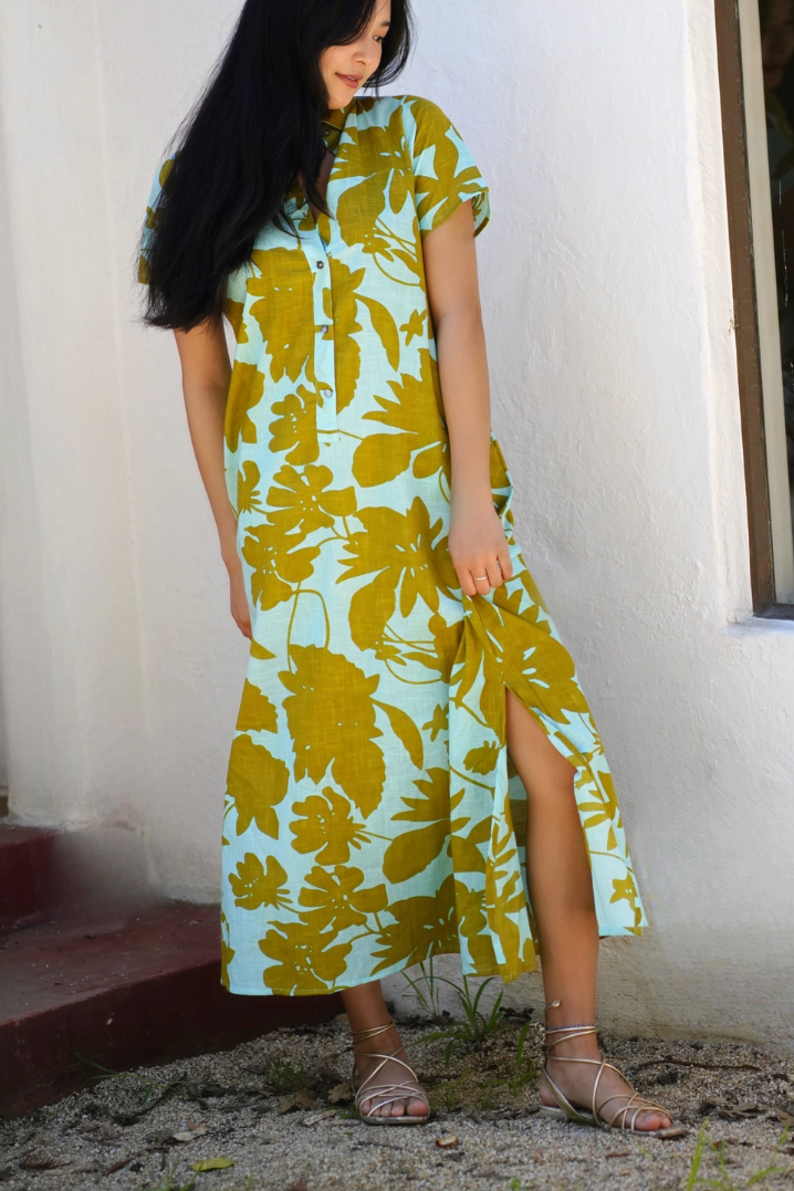 A woman wearing a short-sleeve midi dress with a light blue and olive green tropical leaf pattern, standing outdoors against a white wall.