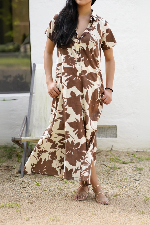 A woman wearing a short-sleeve button-front maxi dress with a large brown floral print on a cream background, standing outdoors in front of a white wall.