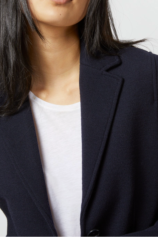 A close-up of a model wearing a dark navy blue blazer over a white crew-neck t-shirt, cropped from the lower face to the waist against a light gray background.