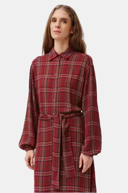 A woman modeling a long-sleeve red and dark green plaid shirt dress with a matching tie belt, set against a plain light gray studio background.