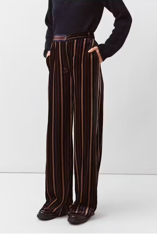 A model wearing high-waisted, wide-leg trousers in a dark brown velvet fabric with vertical stripes in black, beige, and muted orange, styled with a black ribbed sweater and dark loafers against a plain white studio background.