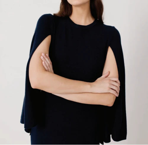 A woman modeling a black sleeveless top with wide draped cap sleeves and arm cutouts, posed against a plain light gray studio background.