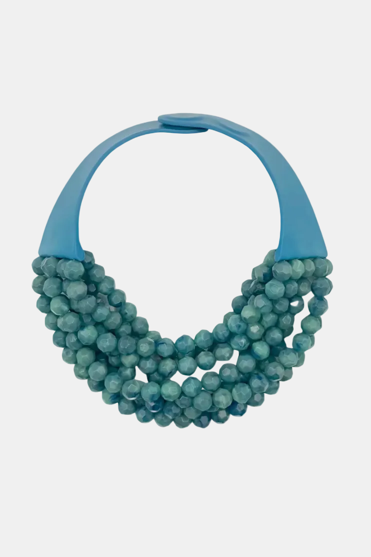 A multi-strand statement necklace featuring textured, faceted round beads in a dusty teal-blue color, attached to a smooth, curved light blue plastic collar, shown against a plain white background.