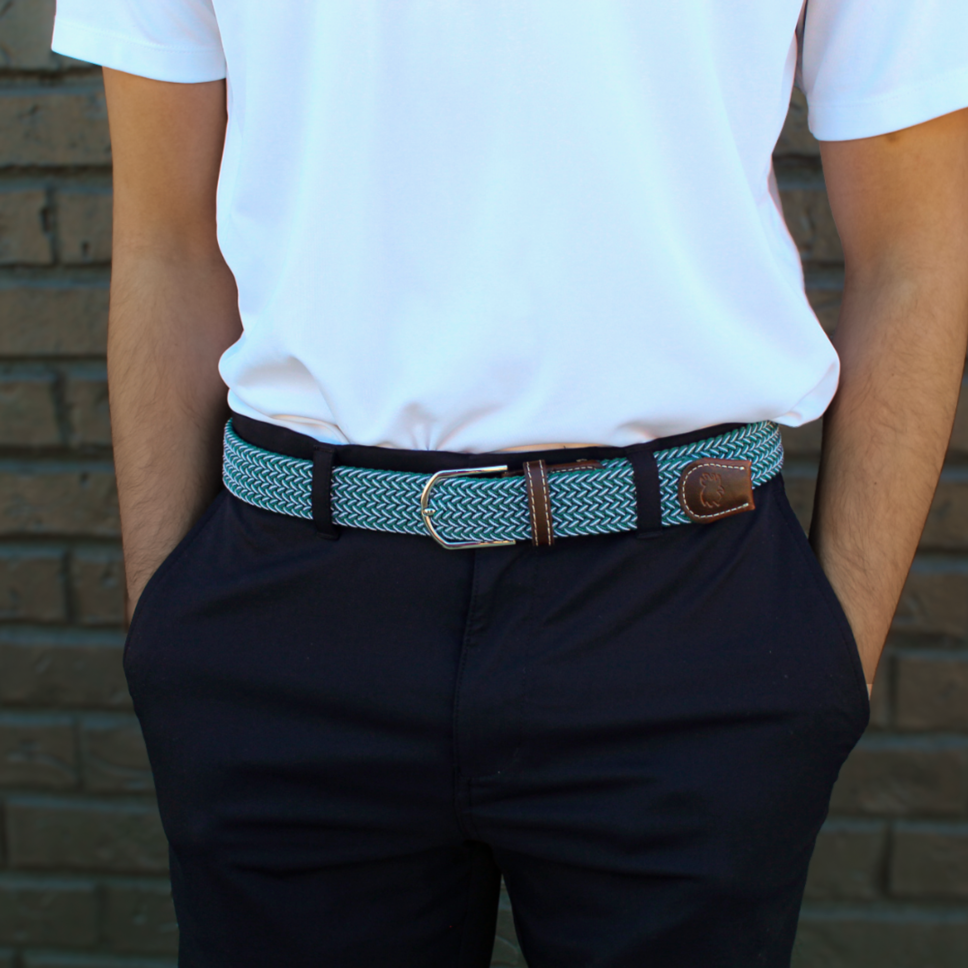 A close-up of a person wearing a patterned woven belt with a silver buckle and brown leather accents, styled with a white polo shirt and dark trousers against a brick wall.