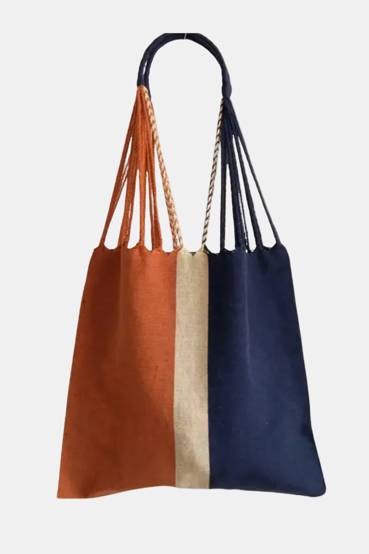 A large fabric tote bag with a vertical three-color design: rust orange on the left, natural beige in the center, and navy blue on the right. It features a single dark handle with mixed rope and fabric attachment methods, shown against a plain white background.