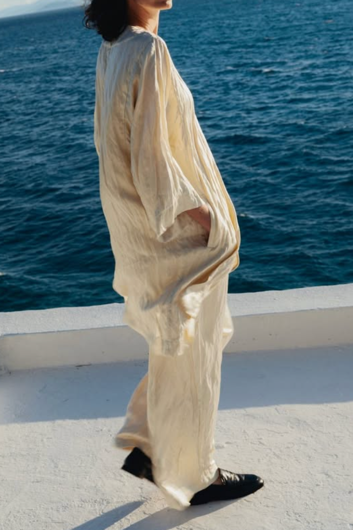 A woman stands on a white balcony overlooking a deep blue ocean, wearing a flowing, loose-fitting cream-colored linen outfit with black shoes.